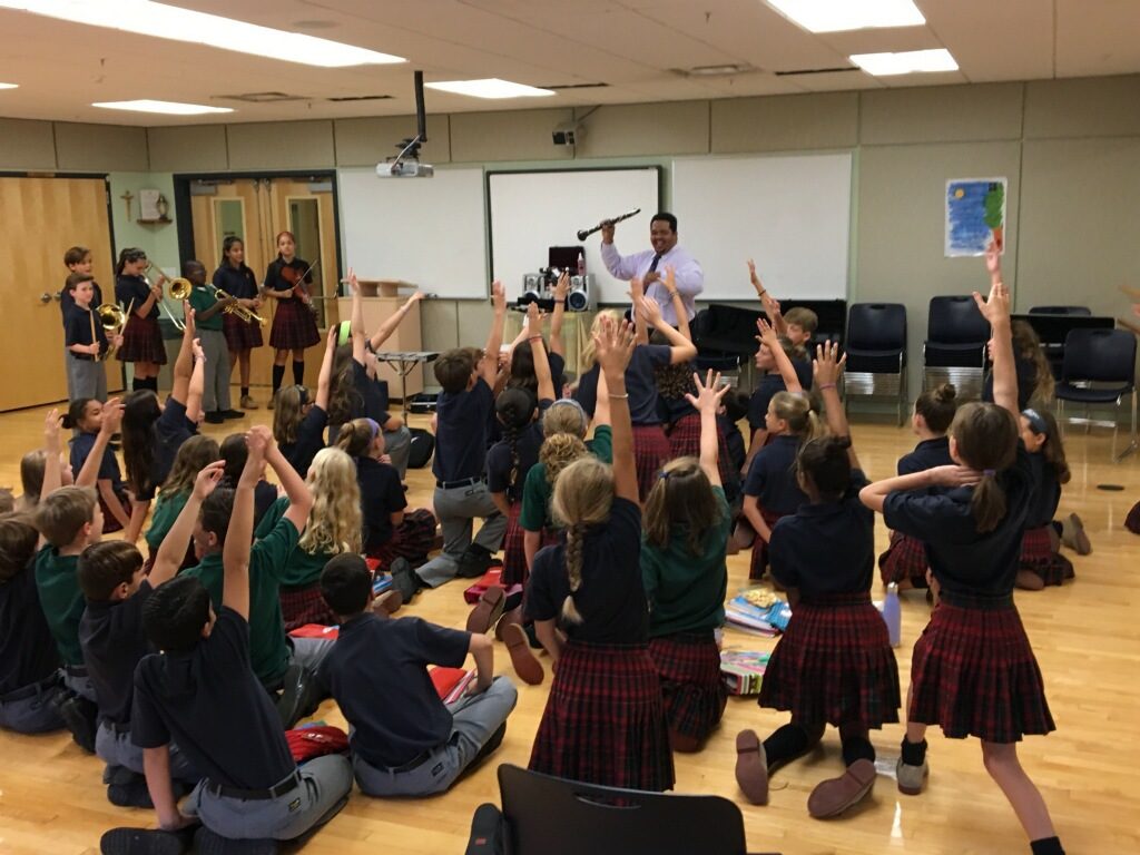 Teacher holding a clarinet in front of a classroom of students sitting on the floor.