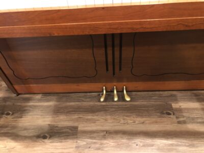 View of the pedals of a Yamaha M405 upright, to show the cherry finish of the instrument and the polished gold finish of the pedals.