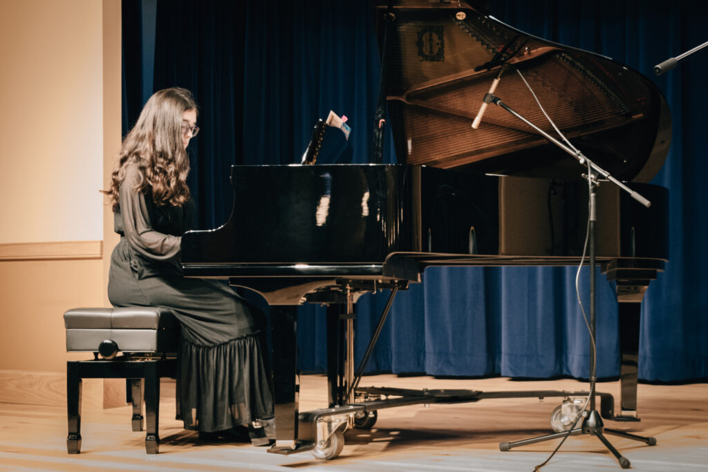 Piano student in a black dress performing at recital on a black grand piano.
